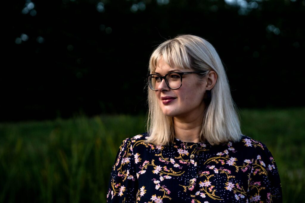 Blonde woman wearing glasses and a floral top looking to the side with a green outdoors background