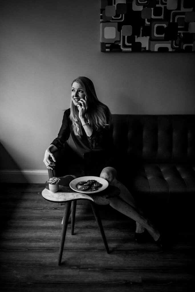 black & white edit of a white woman with blonde hair sitting on an office sofa whilst chatting on a mobile phone looking out of the window. There's a plate of biscuits on a small table in front of her.