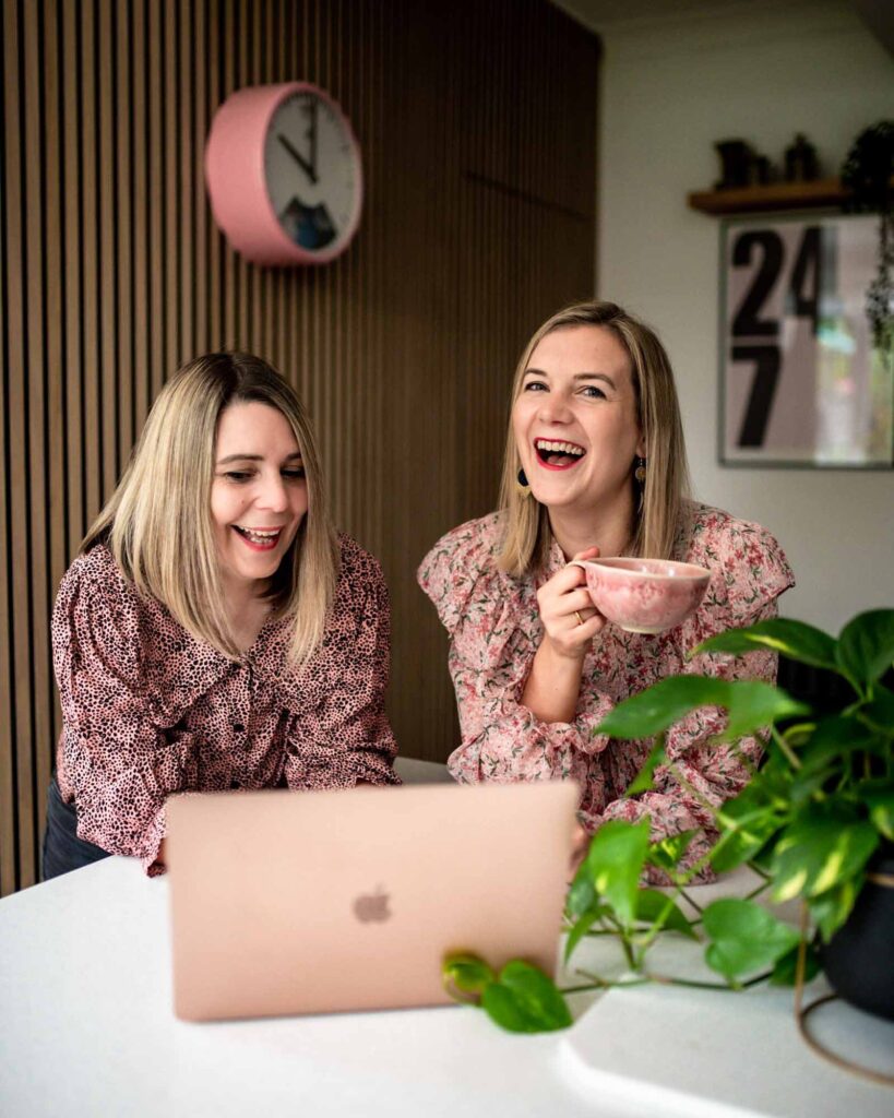 Blonde sisters laughing in a kitchen with a pink mug and a rose gold Mac laptop
