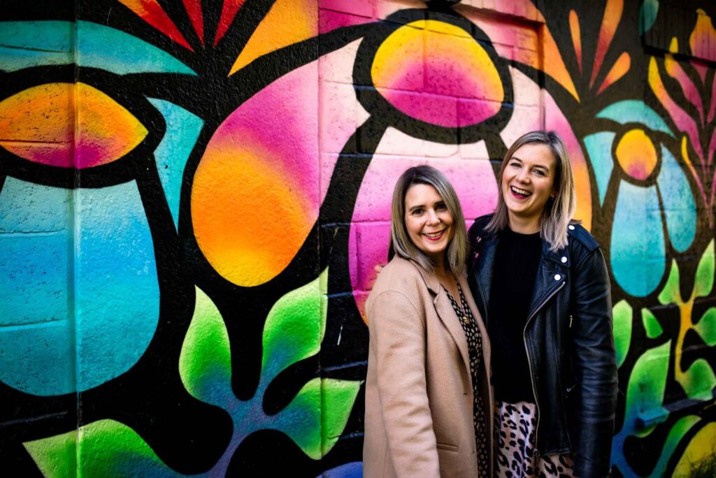 Two white women with blonde hair posing in front of a colourful mural while smiling