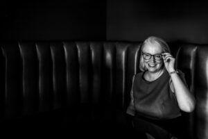 B&W edit of a white woman holding her glasses, sitting in a booth table in a hotel in Brighton