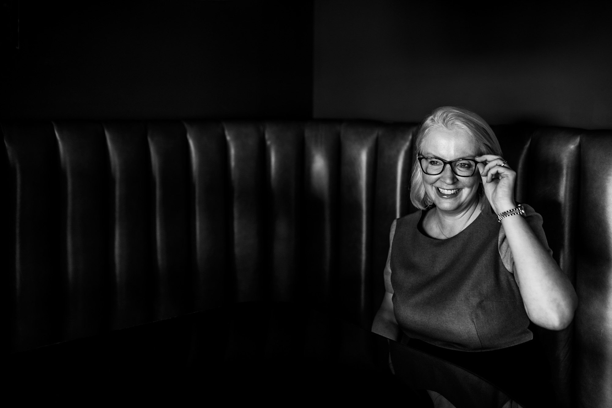 B&W edit of a white woman holding her glasses, sitting in a booth table in a hotel in Brighton