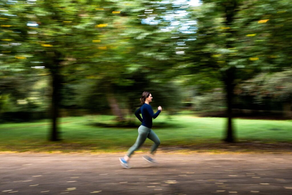 A white woman with brown hair running through a blurred green park in London. She is wearing green leggings and a navy top.