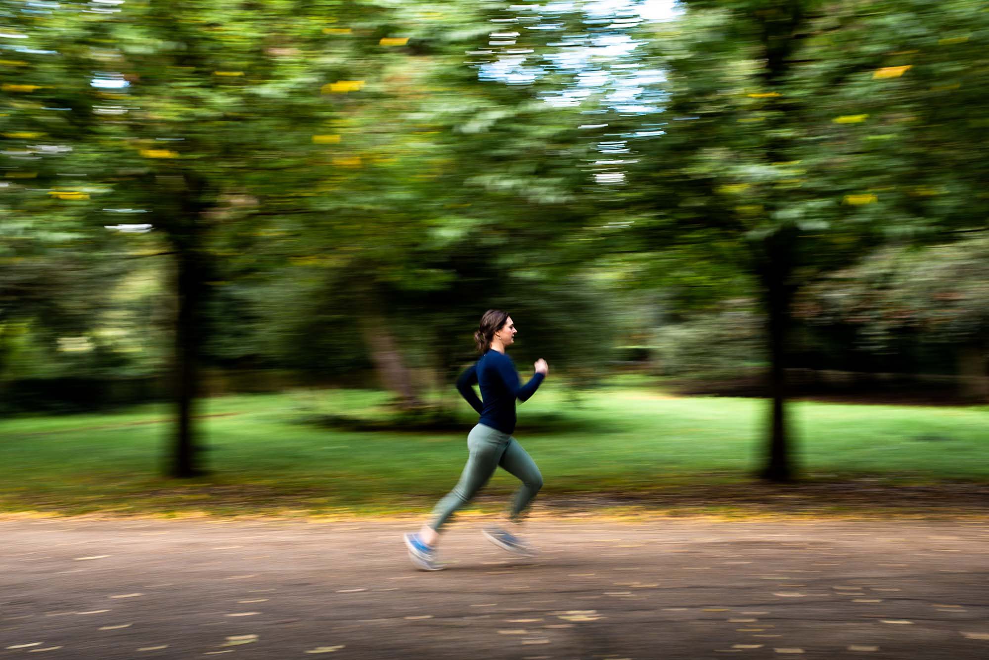 A white woman with brown hair running through a blurred green park in London. She is wearing green leggings and a navy top.