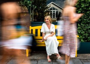 White woman in a white dress sitting on a yellow bench with people rushing past in a blur.