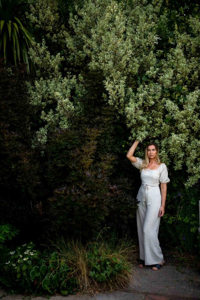 A white blond woman in a white jumpsuit posing surrounded by trees in a park