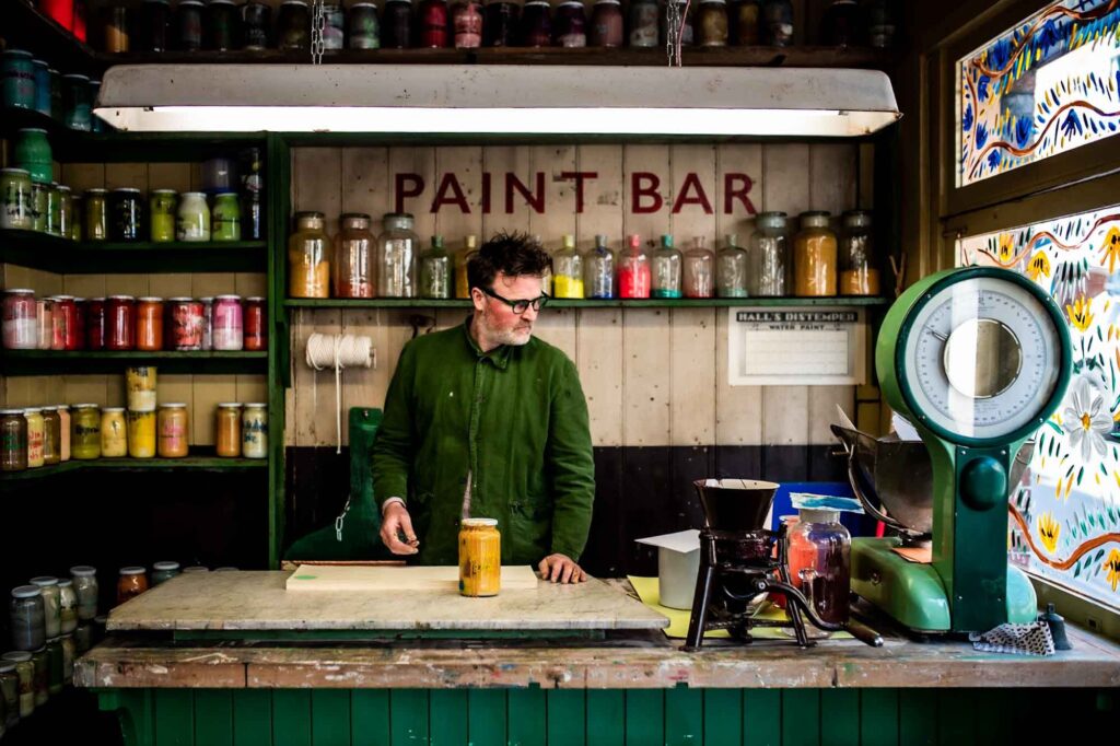 White man behind the counter of a "Paint Bar" in a colourful shop in Lewes, surrounded by vibrant jars of pigment.