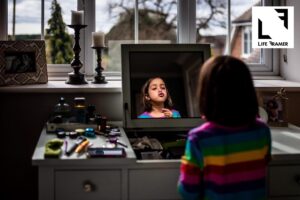 A young girl wearing a rainbow striped dress is sitting at her mother's dressing table mirror in front of a window, surrounded by make-up and pursing her lips in the reflection whilst holding pink lipstick. There is a "Life Framer" logo in the corner.