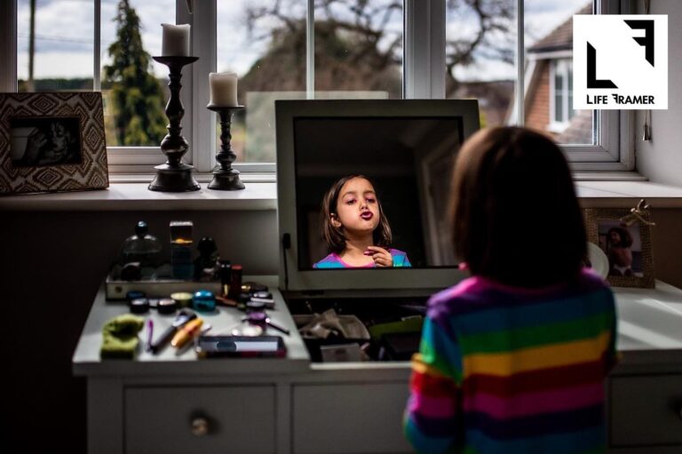 A young girl wearing a rainbow striped dress is sitting at her mother's dressing table mirror in front of a window, surrounded by make-up and pursing her lips in the reflection whilst holding pink lipstick. There is a "Life Framer" logo in the corner.