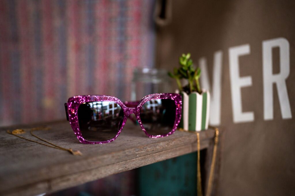 Product photography showing pink sparkly glitter sunglasses on a wooden shelf
