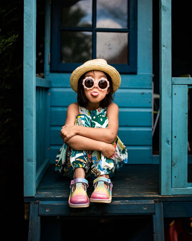 A mixed race girl wearing a straw hat, funky floral pink sunglasses, a tropical print jumpsuit and rainbow Kickers shoes for their brand campaign. She's sitting on a blue playhouse porch with her arms around her bent legs and sticking her tongue out.