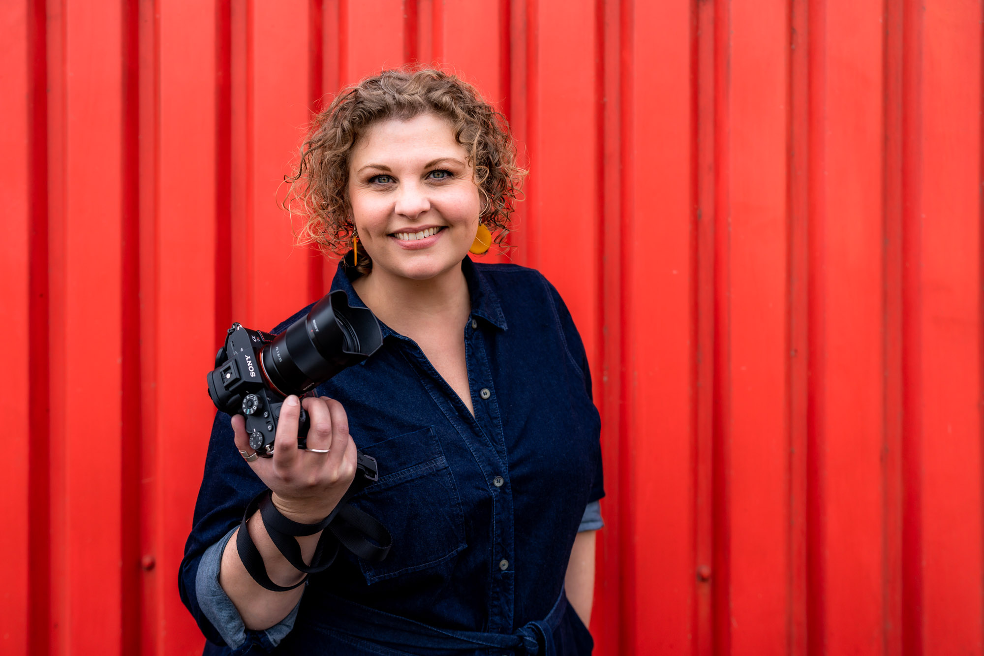 A white female photographer with light brown curly hair poses in a denim dress for a headshot with a red backdrop