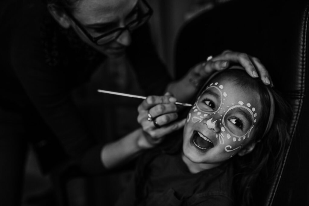 Make-up artists painting the face of a young girl with a sugar skull pattern in black & white