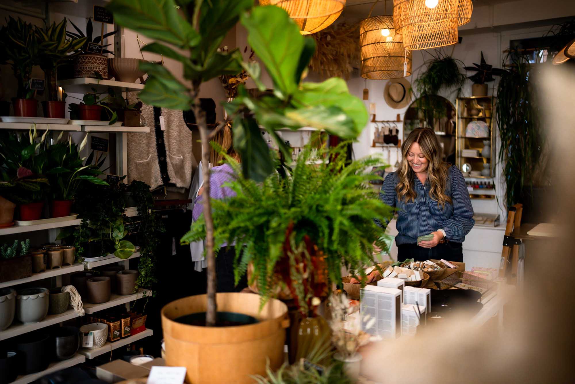 A white woman with blonde hair wearing a blue shirt. She's organising her lifestyle shop, From Victoria in Lewes, surrounded by greenery and beautiful gifts.