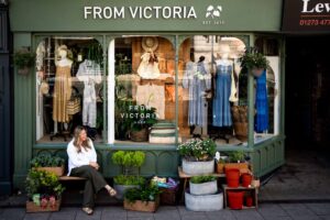 Victoria, a white woman with long blonde hair in a white shirt and green trousers, sitting on a bench laughing outside her green-fronted shop, From Victoria, with summer dresses in the window and plants on the pavement.
