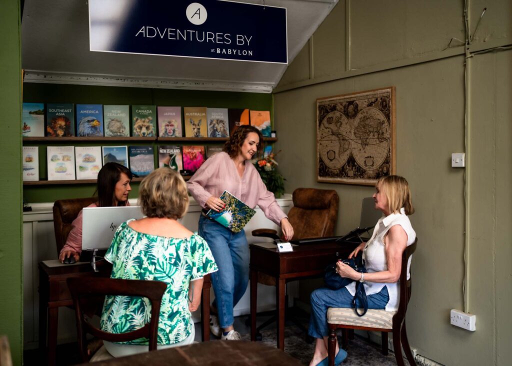 4 women sitting together at a travel agent hub office with sage green walls and travel brochures in the background.