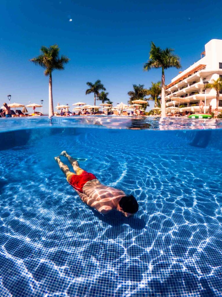 Man in red trunks swimming underwater in a blue swimming pool with palm trees in the background