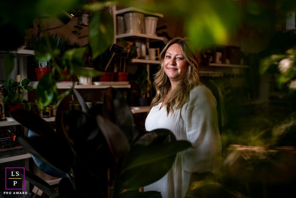 Victoria, a white woman with long blonde hair in a white top, smiling softly at the camera, framed by dark plant leaves in her shop, with an LPA gold medal logo.