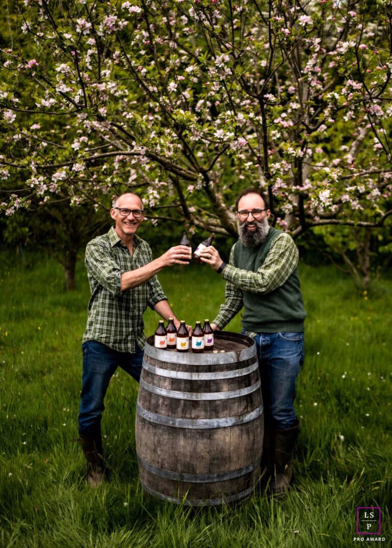 Two men wearing green tartan shirts and blue jeans are saying cheers as they clink their Bignose & Beardy cider bottles during their brand photoshoot. They're smiling at the camera and leaning on an old wooden barrel situated in a green orchard in Sussex, with blossoming apple trees around them. There is a "Pro Award" symbol in the corner.