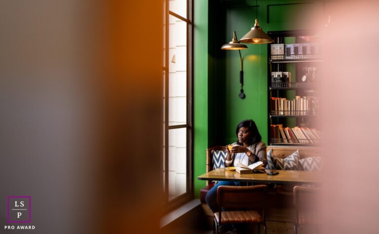 A black female doctor, wearing a brown blazer, white t-shirt and blue jeans, enjoying a cup of coffee in a yellow mug. She's inside a London café with a green background, surrounded by lots of books and looking out of a large window. There is a "Pro Award" symbol in the corner.