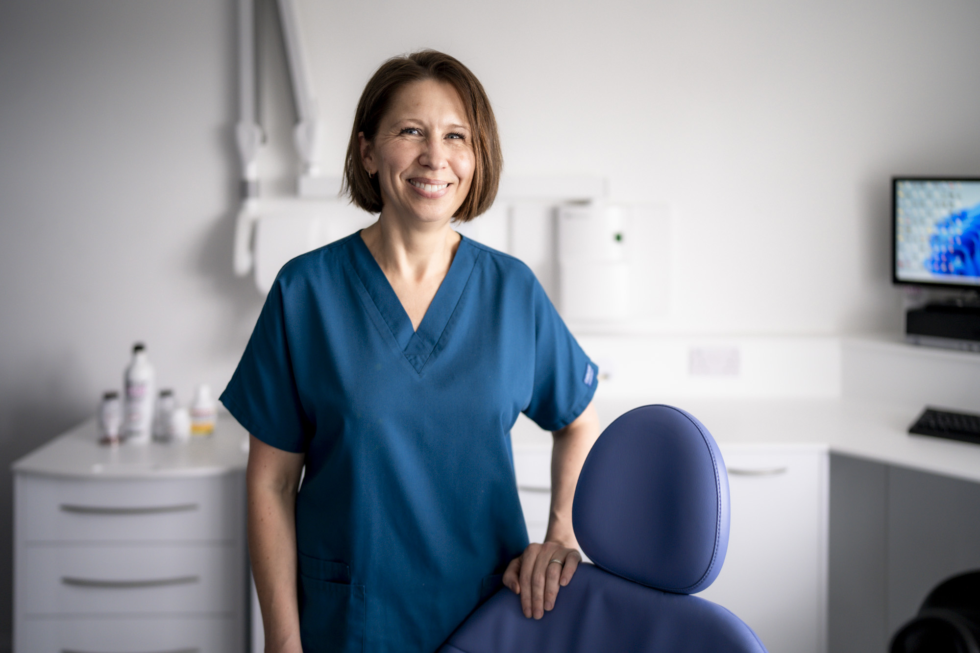 Kate, a smiling hygienist with shoulder-length dark hair in teal scrubs, standing beside a blue dental chair in a bright white surgery at Durban Dental Centre, Crowborough, East Sussex.