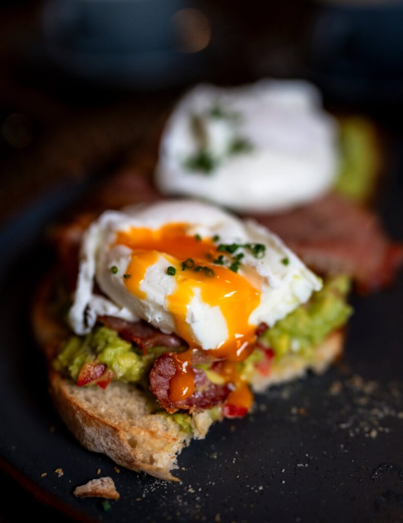Close-up of a poached egg with a runny orange yolk on toasted sourdough with smashed avocado and chorizo, sprinkled with chives, on a dark plate.