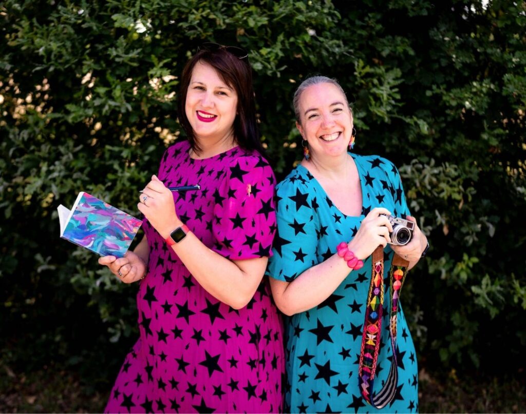 Two white women in Scamp & Dude star print colourful pink & teal dresses standing back to back, one holding a notebook and one has a camera.
