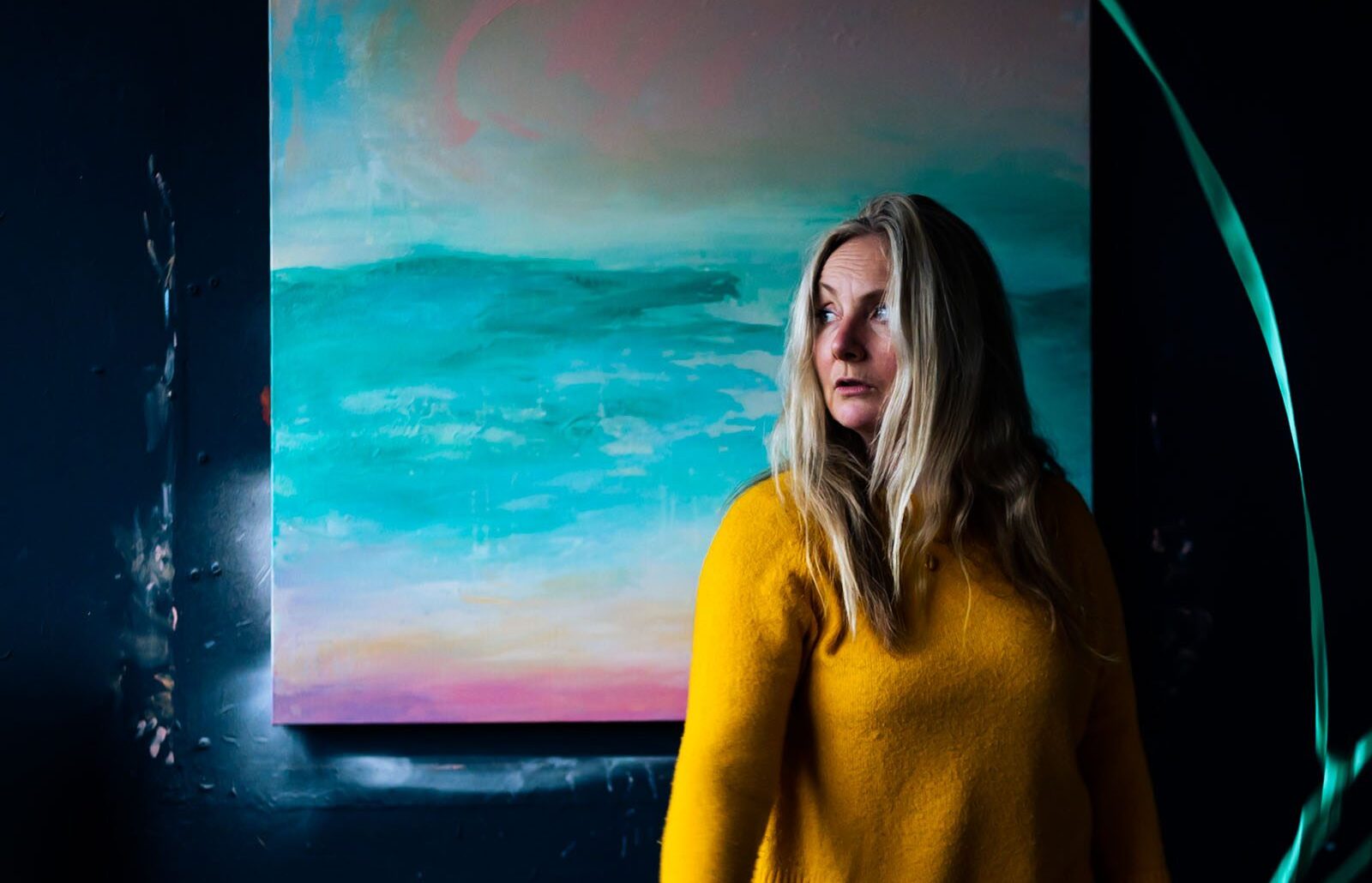 Antonia, a white woman with long blonde hair in a mustard yellow jumper, gazing to the side in front of a large teal and pink abstract seascape painting.