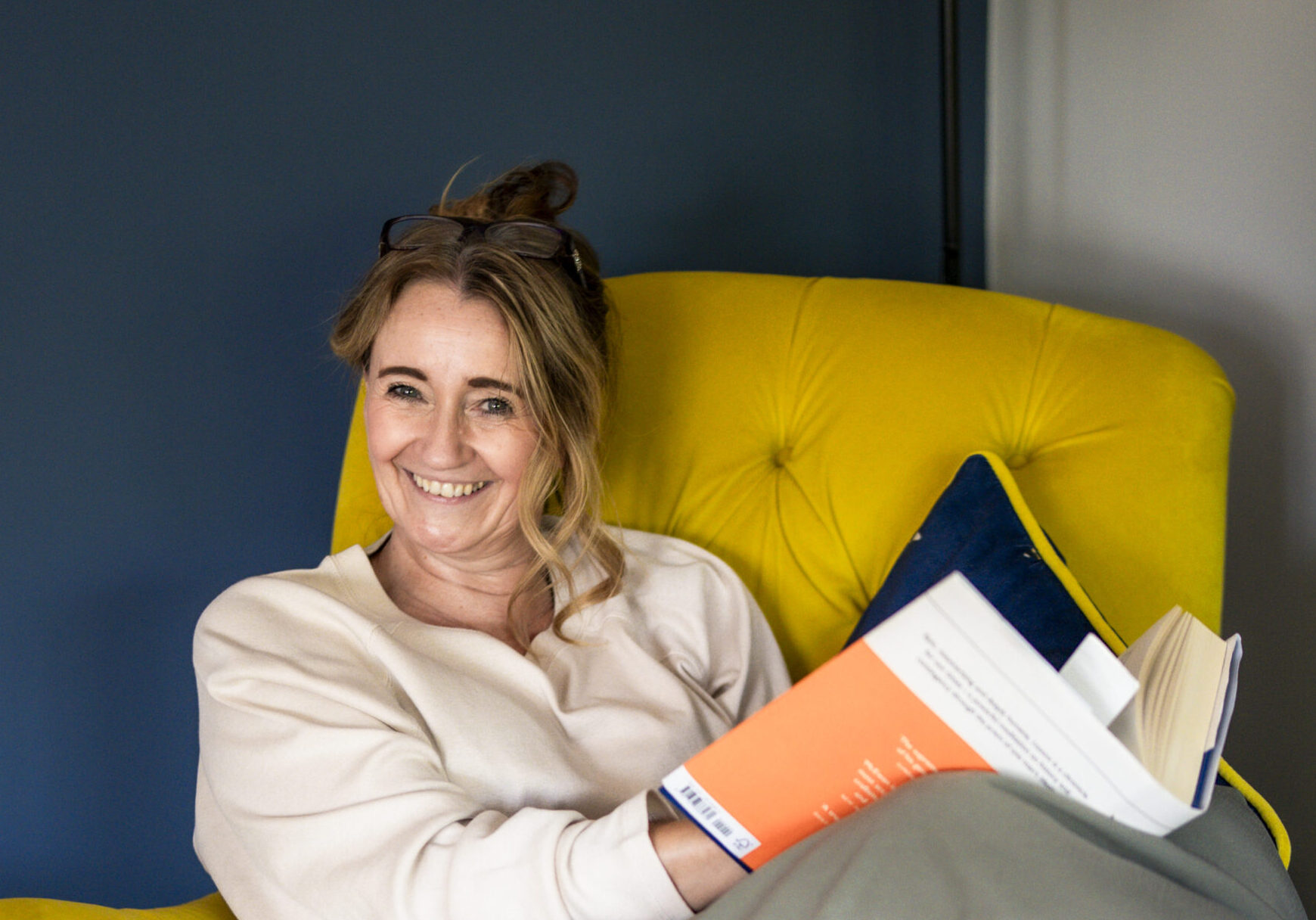 A smiling woman with wavy, light brown hair pinned up and glasses on her head, wearing a cream jumper and reading a book in a yellow velvet armchair against a navy wall, during her photoshoot as a GDPR consultant