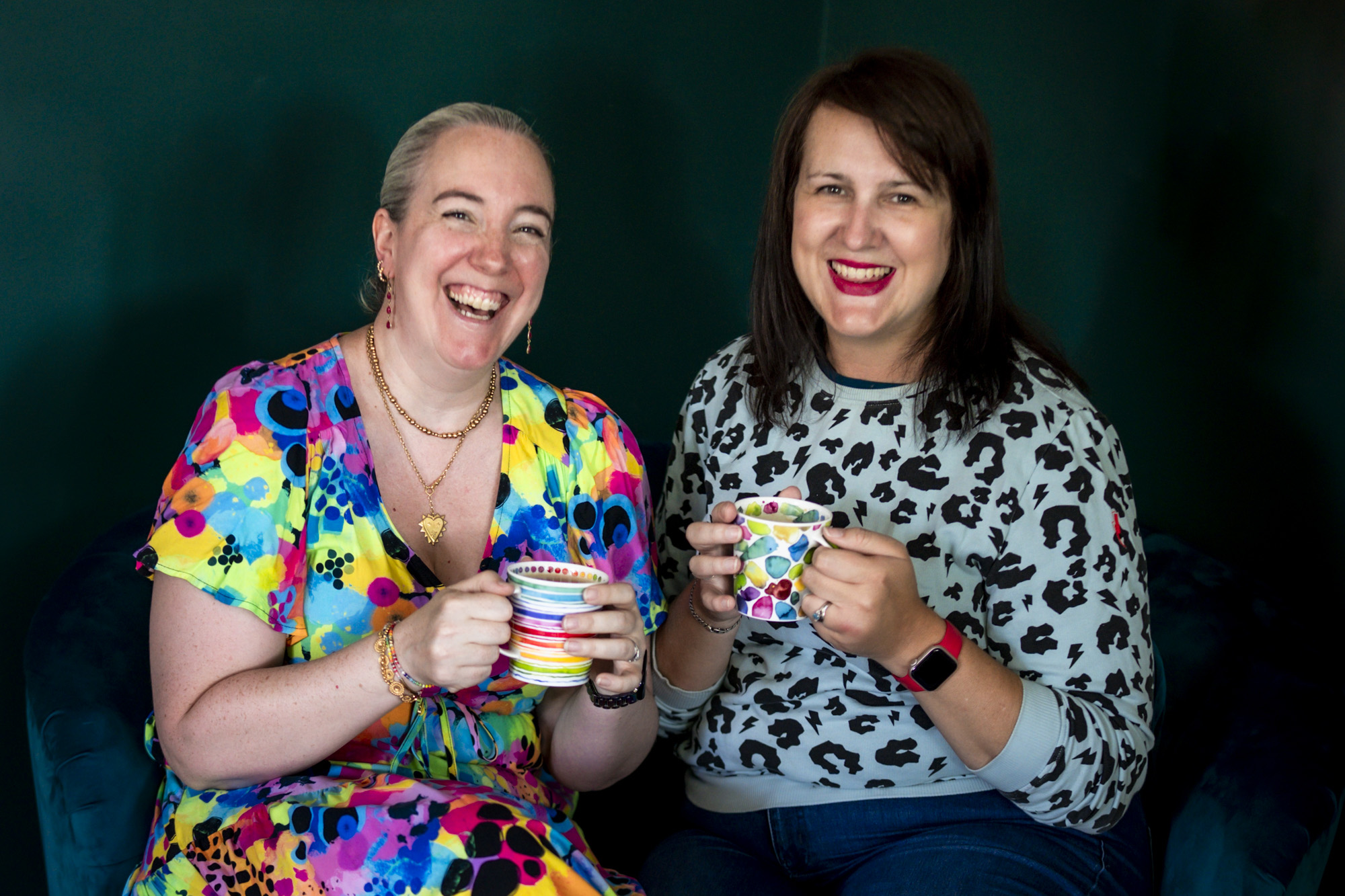 Bex Maini and Jo Martin laughing together against a dark background, each holding a mug - Bex, on the left, is a white woman with short grey hair in a bright, multicoloured patterned dress, and Jo, on the right, is a white woman with dark hair in a black and white animal-print top.