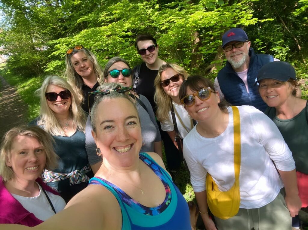 A selfie of eight smiling people on a sunny woodland walk under bright green trees. At the front is a white woman with grey hair pulled back, sunglasses on her head and a blue patterned top, with a group of women in sunglasses and a bearded man in a cap and navy gilet behind her.