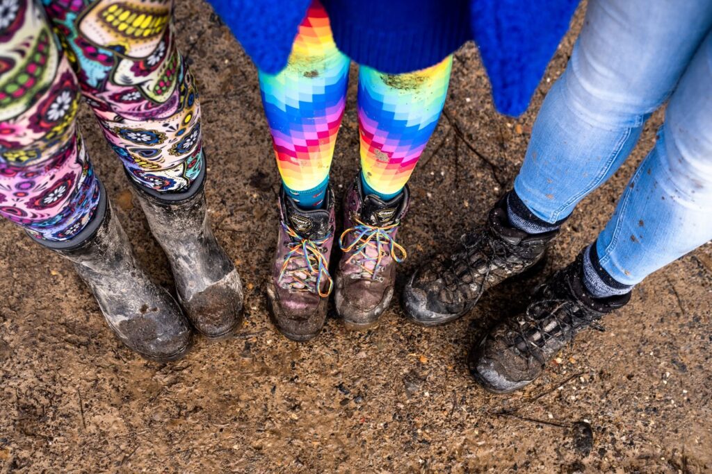 Looking down at three pairs of muddy walking boots and wellies on a wet path, one in colourful patterned leggings, one in rainbow zigzag tights with rainbow laces, and one in light blue jeans.