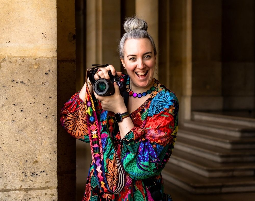 A happy, smiling white woman in a multi-coloured dress is holding her camera up to take photos in Paris