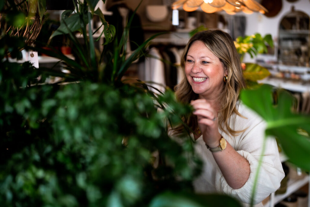 Victoria, a white woman with long blonde hair and gold earrings, smiling broadly while tending a leafy green plant in her shop.