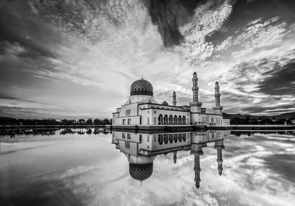 A black and white photo of a domed mosque with minarets, reflected in still water beneath a dramatic cloudy sky in Borneo.
