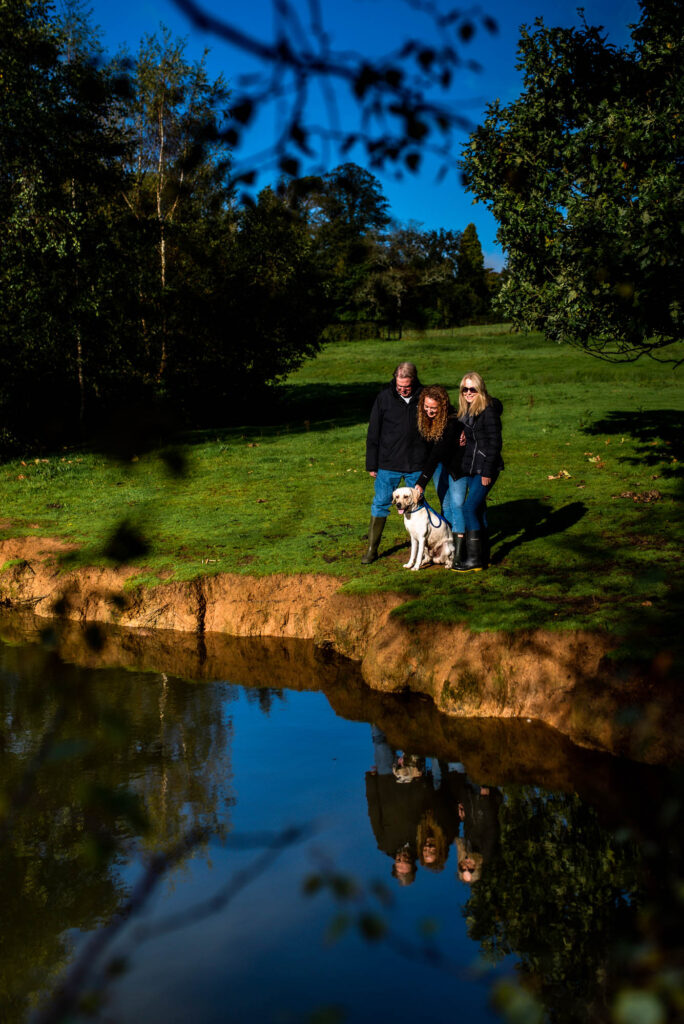 A grey-haired white man and two white women, one with curly auburn hair and one blonde in sunglasses, stand with a yellow Labrador by a pond, reflected in the water
