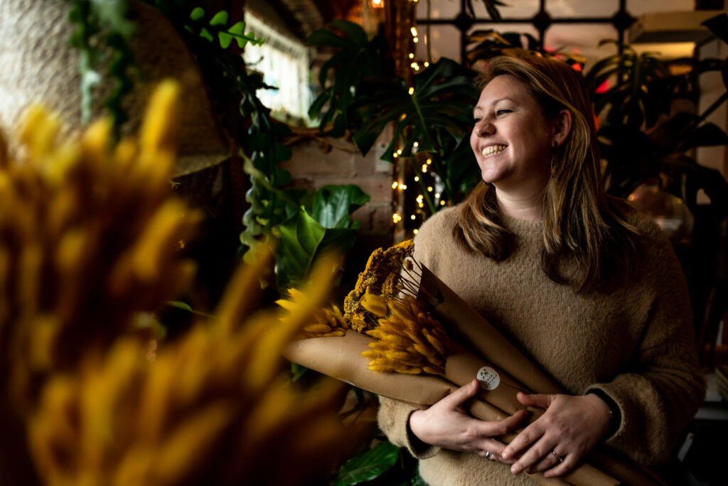 Victoria, a white woman with shoulder-length blonde hair and a fluffy beige jumper, laughing as she looks out of a window, holding paper-wrapped bunches of yellow dried flowers and grasses, with fairy lights and plants behind her and yellow grasses blurred in the foreground.