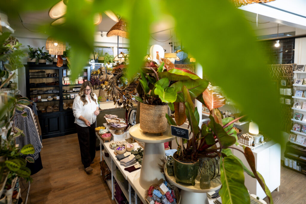 Looking down through soft green leaves into a lifestyle shop, where a white woman with long blonde hair and glasses in a white linen shirt arranges a table of soaps and socks, surrounded by large potted plants, woven pendant lamps, a dried flower wreath and shelves of homewares and cards.