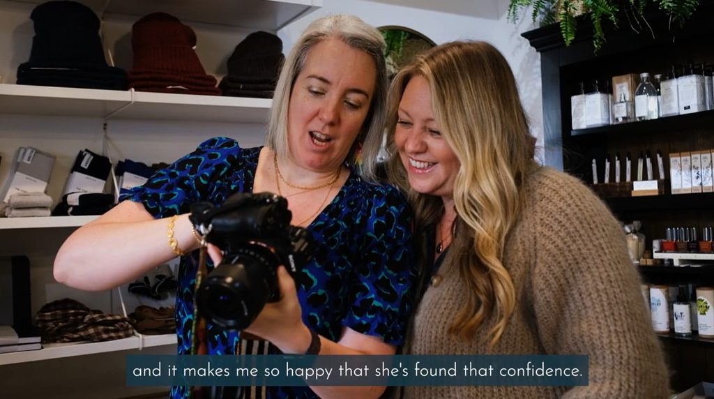 A still from a brand film: two women looking at the back of a camera together in a shop, one with short grey hair in a blue patterned dress and one with long blonde hair in a knitted cardigan, both smiling. The subtitle reads “and it makes me so happy that she’s found that confidence.”