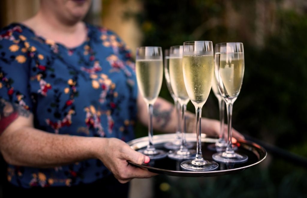 A woman in a blue floral top holding a silver tray of champagne flutes at an Uckfield Chamber of Commerce networking event at Horsted Place Hotel.