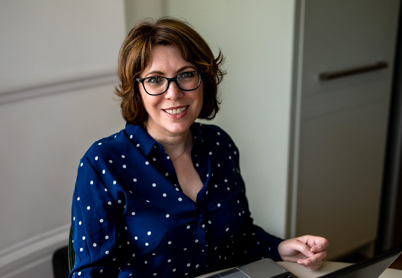 Jane Griffin of Positive Story PR, a smiling white woman with short brown hair and glasses in a navy polka dot blouse, sitting at a desk with a laptop.
