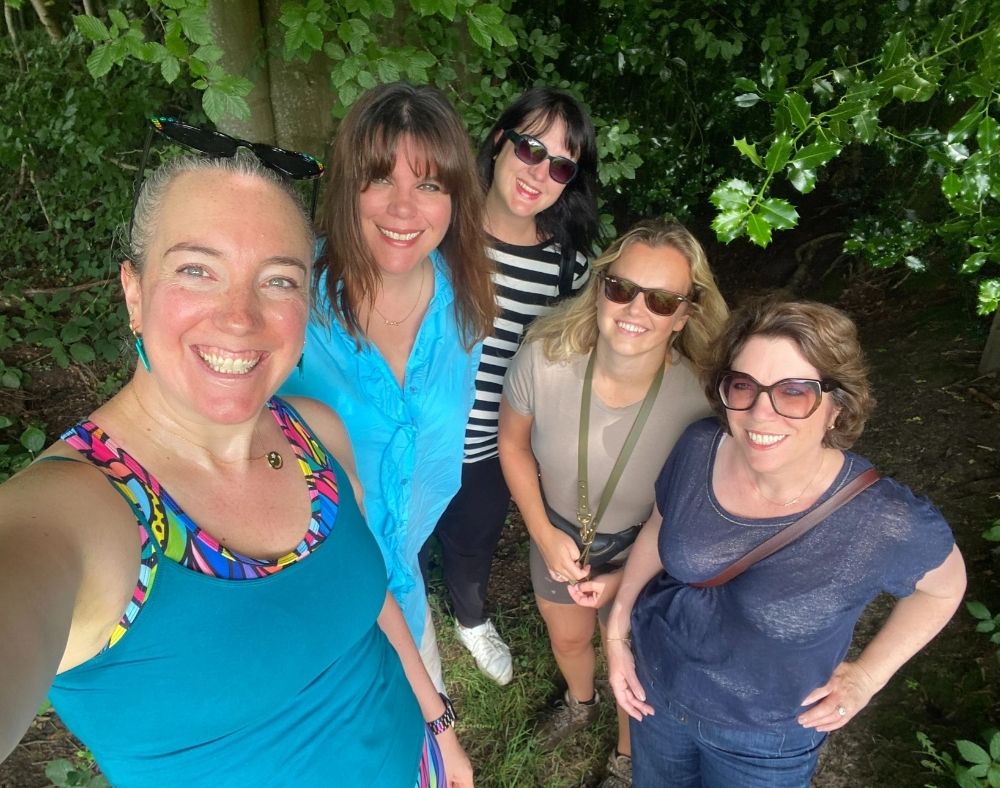 A selfie of five smiling women on a woodland walk surrounded by green leaves. At the front is a white woman with grey hair pulled back, sunglasses on her head and a teal top over a rainbow-patterned top. Behind her are a woman with long brown hair and a fringe in a turquoise shirt, a woman with dark hair and sunglasses in a black and white striped top, a woman with blonde hair and sunglasses in a beige T-shirt, and a woman with short brown hair and glasses in a navy top.