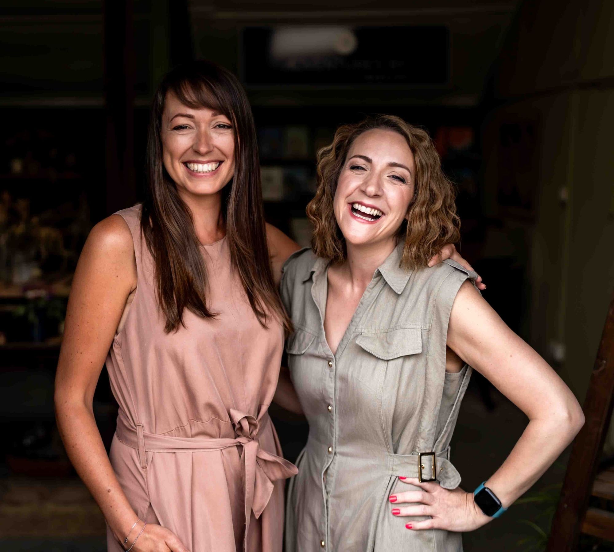 Two white women with brown hair standing in the entrance to a travel agent shop in Sussex. One is in a dusky pink dress and smiling, the other is wearing a khaki green dress and laughing.