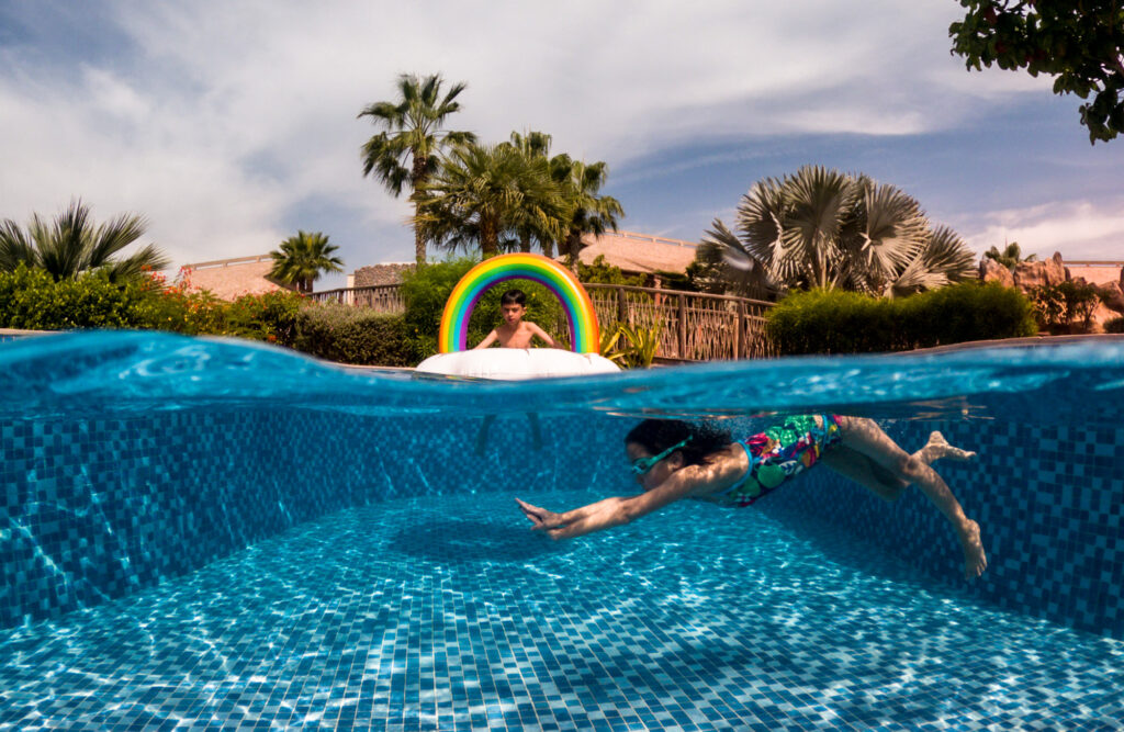 A split-level view of a child swimming underwater in a bright blue pool in Dubai, with another child floating on a rainbow inflatable above and palm trees behind.